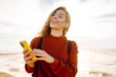 Gün batımında kumsalda gülümseyen ve akıllı telefonunu kullanan genç bir kadın. Altın güneş ışığı saçlarını ve çevresini aydınlatıyor. Bloglama, iletişim konsepti.