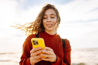 Gün batımında kumsalda gülümseyen ve akıllı telefonunu kullanan genç bir kadın. Altın güneş ışığı saçlarını ve çevresini aydınlatıyor. Bloglama, iletişim konsepti.