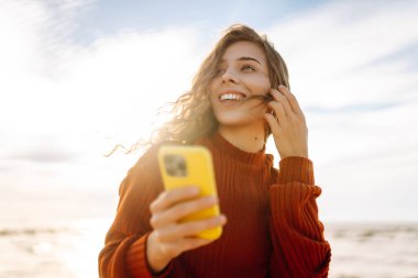 Gün batımında kumsalda gülümseyen ve akıllı telefonunu kullanan genç bir kadın. Altın güneş ışığı saçlarını ve çevresini aydınlatıyor. Bloglama, iletişim konsepti.