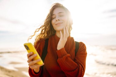 Gün batımında kumsalda gülümseyen ve akıllı telefonunu kullanan genç bir kadın. Altın güneş ışığı saçlarını ve çevresini aydınlatıyor. Bloglama, iletişim konsepti.