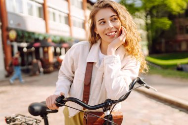 Kıvırcık saçlı genç bir kadın güneşli bir günün tadını çıkarırken etrafı dükkanlarla çevrili güzel bir caddede bisikletiyle poz veriyor. Aktif yaşam tarzı. Eko taşımacılığı.