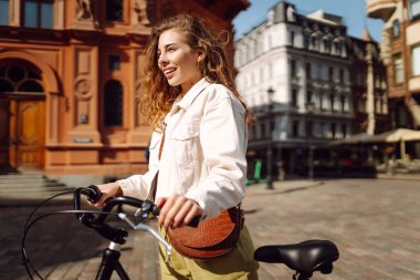 Kıvırcık saçlı genç bir kadın güneşli bir günün tadını çıkarırken etrafı dükkanlarla çevrili güzel bir caddede bisikletiyle poz veriyor. Aktif yaşam tarzı. Eko taşımacılığı.
