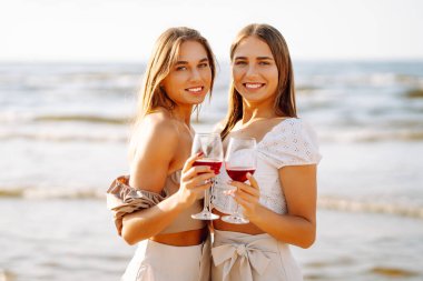 Gün batımında güneşli bir plajda kırmızı şaraptan zevk alan iki kadın, deniz kenarında arkadaşlığı ve rahatlığı kutluyorlar. Moda tarzı. Piknik konsepti.