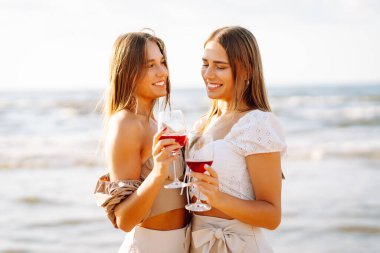Gün batımında güneşli bir plajda kırmızı şaraptan zevk alan iki kadın, deniz kenarında arkadaşlığı ve rahatlığı kutluyorlar. Moda tarzı. Piknik konsepti.