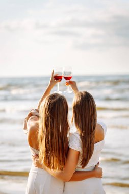 Gün batımında güneşli bir plajda kırmızı şaraptan zevk alan iki kadın, deniz kenarında arkadaşlığı ve rahatlığı kutluyorlar. Moda tarzı. Piknik konsepti.