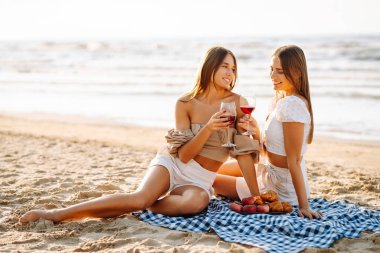 Gün batımında güneşli bir plajda kırmızı şaraptan zevk alan iki kadın, deniz kenarında arkadaşlığı ve rahatlığı kutluyorlar. Moda tarzı. Piknik konsepti.