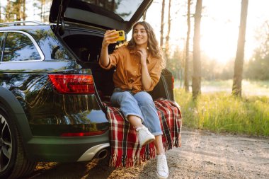 Günbatımında güneşli bir ormanda arabasının bagajında otururken akıllı telefonuyla selfie çeken neşeli bir kadın. Arabayla seyahat etme kavramı, aktif yaşam tarzı..