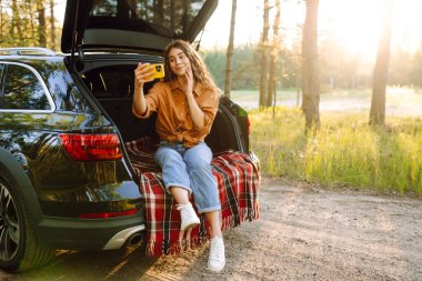 Günbatımında güneşli bir ormanda arabasının bagajında otururken akıllı telefonuyla selfie çeken neşeli bir kadın. Arabayla seyahat etme kavramı, aktif yaşam tarzı..