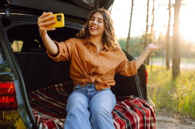 Günbatımında güneşli bir ormanda arabasının bagajında otururken akıllı telefonuyla selfie çeken neşeli bir kadın. Arabayla seyahat etme kavramı, aktif yaşam tarzı..