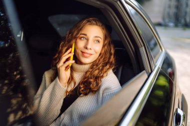 Kıvırcık saçlı genç bir kadın arabada otururken gülümsüyor, akıllı telefonunu tutuyor. İş, teknoloji, seyahat, online iletişim kavramı. Taksi..