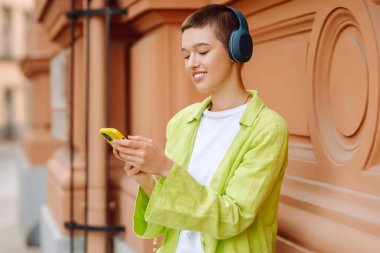 Parlak yeşil ceketli genç bir kadın, şehir merkezinde akıllı telefonunu kullanırken kulaklıkla müzik dinlemekten hoşlanıyor