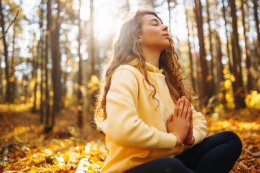 Huzurlu bir kadın, sonbahar yapraklarıyla çevrili güneşli bir ormanda yoga meditasyonu yapıyor. Yaşam tarzı ve meditasyon konsepti.