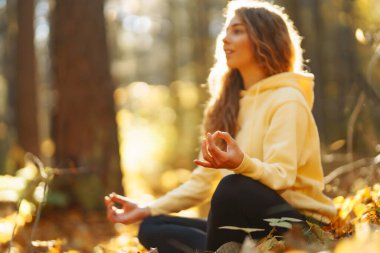 Huzurlu bir kadın, sonbahar yapraklarıyla çevrili güneşli bir ormanda yoga meditasyonu yapıyor. Yaşam tarzı ve meditasyon konsepti.