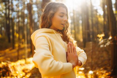 Huzurlu bir kadın, sonbahar yapraklarıyla çevrili güneşli bir ormanda yoga meditasyonu yapıyor. Yaşam tarzı ve meditasyon konsepti.