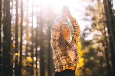 Huzurlu bir kadın, sonbahar yapraklarıyla çevrili güneşli bir ormanda yoga meditasyonu yapıyor. Yaşam tarzı ve meditasyon konsepti.