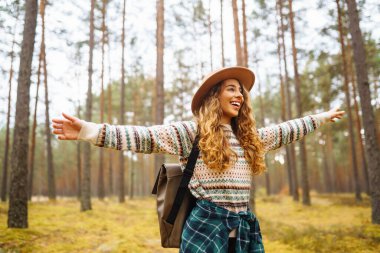 Sonbaharda sakin bir orman yolunu keşfeden genç bir gezgin kadın rahat bir süveter ve geniş kenarlı sırt çantalı bir şapka giyiyor. Seyahat, doğa konsepti..