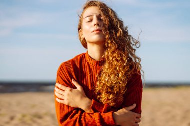 Kıvırcık saçlı genç bir kadın güneşli kumsalın tadını çıkarıyor. Açık bir günün sıcaklığında gülümsüyor ve güneşleniyor. Bahar geldi. Seyahat, hafta sonu, rahatlama ve yaşam tarzı konsepti.