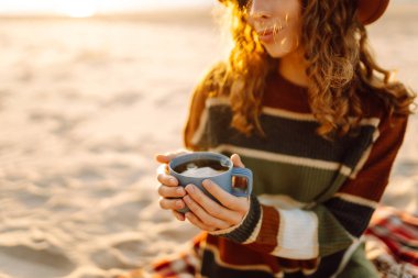 Genç bir kadın kumlu sahilde oturmuş elinde sıcak bir içkiyle güzel günbatımının tadını çıkarıyor. Renkli bir süveter ve şapka giyiyor, sakin atmosferi kucaklıyor. Piknik, hafta sonu, rahatlama ve yaşam tarzı