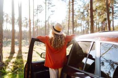Ormanda arabasının yanında güneşli bir günün tadını çıkaran bir kadın ağaçlar arasında doğanın güzelliğini kucaklıyor ve gülümsüyor. Arabayla seyahat etme kavramı, aktif yaşam tarzı..