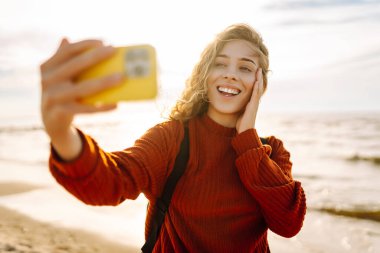 Genç bir kadın, arka planda okyanus dalgalarıyla güneşli bir sahilde selfie çekiyor. Selfie zamanı. Soğuk rüzgarlı hava. Açık havada..