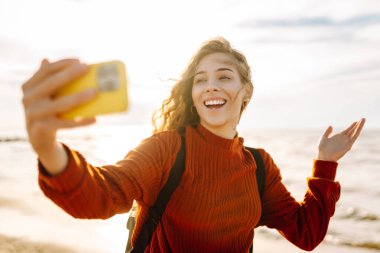 Genç bir kadın, arka planda okyanus dalgalarıyla güneşli bir sahilde selfie çekiyor. Selfie zamanı. Soğuk rüzgarlı hava. Açık havada..