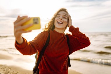 Genç bir kadın, arka planda okyanus dalgalarıyla güneşli bir sahilde selfie çekiyor. Selfie zamanı. Soğuk rüzgarlı hava. Açık havada..