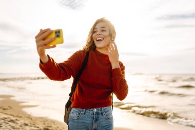 Genç bir kadın, arka planda okyanus dalgalarıyla güneşli bir sahilde selfie çekiyor. Selfie zamanı. Soğuk rüzgarlı hava. Açık havada..