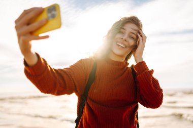 Genç bir kadın, arka planda okyanus dalgalarıyla güneşli bir sahilde selfie çekiyor. Selfie zamanı. Soğuk rüzgarlı hava. Açık havada..