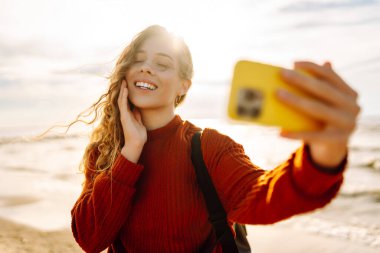 Genç bir kadın, arka planda okyanus dalgalarıyla güneşli bir sahilde selfie çekiyor. Selfie zamanı. Soğuk rüzgarlı hava. Açık havada..
