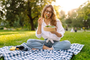 Parkta güneşli bir günde yeşil çimlerle çevrili bir piknik battaniyesinde oturan bir kadın. Kavramsal piknik. Vejetaryen.