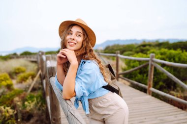 Mutlu genç bir kadın kumsalın yanındaki tahta kaldırımda duruyor, hasır şapka ve mavi gömlek giyiyor, güneş ve okyanus esintisi içinde sırılsıklam. Yaşam tarzı, tatil, seyahat, doğa, aktif yaşam.