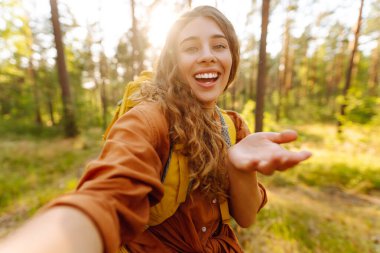 Selfie zamanı! Genç bir kadın, bereketli bir ormanda güneşli bir yürüyüşün keyfini çıkarıyor. Sıcak bir günde, doğal ortamında sırt çantasıyla. Özçekim doğada.