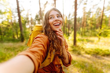 Selfie zamanı! Genç bir kadın, bereketli bir ormanda güneşli bir yürüyüşün keyfini çıkarıyor. Sıcak bir günde, doğal ortamında sırt çantasıyla. Özçekim doğada.