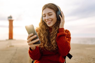 Deniz fenerinin bulutlu bir gününde, deniz kenarında müzik dinleyen genç bir kadın. Seyahat, turizm, teknoloji, blog konsepti.