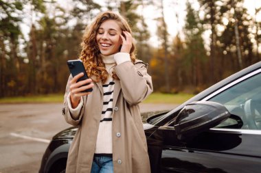 Orman manzaralı bir yerde park halindeki bir arabanın yanında akıllı telefonunu kullanarak neşeli bir anın tadını çıkaran genç bir kadın. Yaşam tarzı, seyahat, turizm, aktif yaşam.
