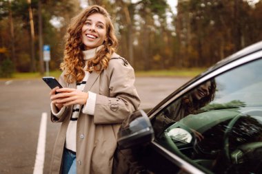 Orman manzaralı bir yerde park halindeki bir arabanın yanında akıllı telefonunu kullanarak neşeli bir anın tadını çıkaran genç bir kadın. Yaşam tarzı, seyahat, turizm, aktif yaşam.