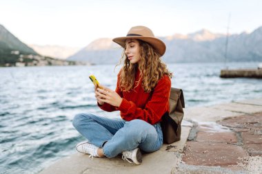 Güneşli bir günde, deniz kenarında manzaralı bir dağ kasabasında telefonunu kullanan genç bir kadın. Tatillerde turist. Yaşam tarzı, seyahat, doğa, aktif yaşam.