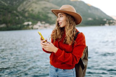 Güneşli bir günde, deniz kenarında manzaralı bir dağ kasabasında telefonunu kullanan genç bir kadın. Tatillerde turist. Yaşam tarzı, seyahat, doğa, aktif yaşam.