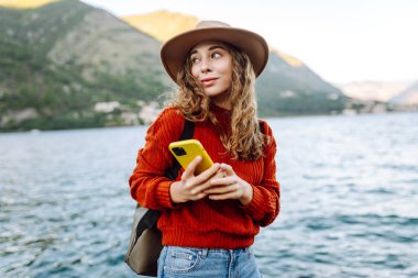 Güneşli bir günde, deniz kenarında manzaralı bir dağ kasabasında telefonunu kullanan genç bir kadın. Tatillerde turist. Yaşam tarzı, seyahat, doğa, aktif yaşam.