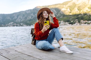 Güneşli bir günde, deniz kenarında manzaralı bir dağ kasabasında telefonunu kullanan genç bir kadın. Tatillerde turist. Yaşam tarzı, seyahat, doğa, aktif yaşam.
