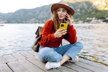 Güneşli bir günde, deniz kenarında manzaralı bir dağ kasabasında telefonunu kullanan genç bir kadın. Tatillerde turist. Yaşam tarzı, seyahat, doğa, aktif yaşam.