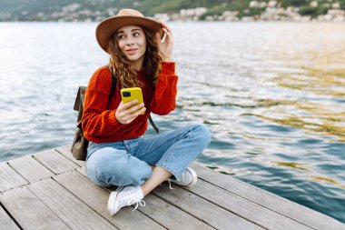 Güneşli bir günde, deniz kenarında manzaralı bir dağ kasabasında telefonunu kullanan genç bir kadın. Tatillerde turist. Yaşam tarzı, seyahat, doğa, aktif yaşam.