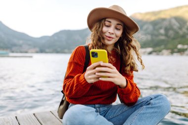Güneşli bir günde, deniz kenarında manzaralı bir dağ kasabasında telefonunu kullanan genç bir kadın. Tatillerde turist. Yaşam tarzı, seyahat, doğa, aktif yaşam.