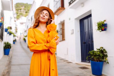 Canlı turuncu elbiseli ve şapkalı genç bir gezgin kadın çiçek saksılarıyla güneşli bir köyde gezintiye çıkıyor. Yaşam tarzı kavramı, tatil, moda, seyahat