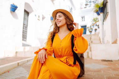 Canlı turuncu elbiseli ve şapkalı genç bir gezgin kadın çiçek saksılarıyla güneşli bir köyde gezintiye çıkıyor. Yaşam tarzı kavramı, tatil, moda, seyahat