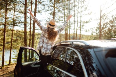 Ekose gömlekli bir kadın göl kenarında huzurlu bir gün batımının keyfini çıkarırken park halindeki arabasının yanında durmuş doğayı kutluyor. Arabayla yolculuk. Yaşam tarzı, seyahat, turizm, doğa konsepti