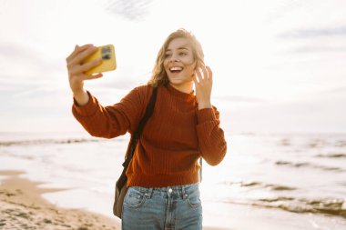 Neşeli genç bir kadın gün batımında güneşli bir sahilde selfie çekerken okyanus dalgalarının tadını çıkarıyor. İnsanlar, tatil, yaşam tarzı, yolculuk konsepti.