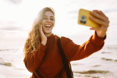 Neşeli genç bir kadın gün batımında güneşli bir sahilde selfie çekerken okyanus dalgalarının tadını çıkarıyor. İnsanlar, tatil, yaşam tarzı, yolculuk konsepti.