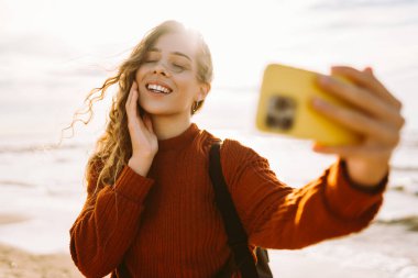 Neşeli genç bir kadın gün batımında güneşli bir sahilde selfie çekerken okyanus dalgalarının tadını çıkarıyor. İnsanlar, tatil, yaşam tarzı, yolculuk konsepti.