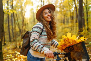 Parkta güneşli bir günde renkli sonbahar yapraklarıyla dolu bir bisikleti iterken gülümseyen genç bir kadın. Aktif turist güneşli havanın tadını çıkarıyor. Hafta sonu konsepti.